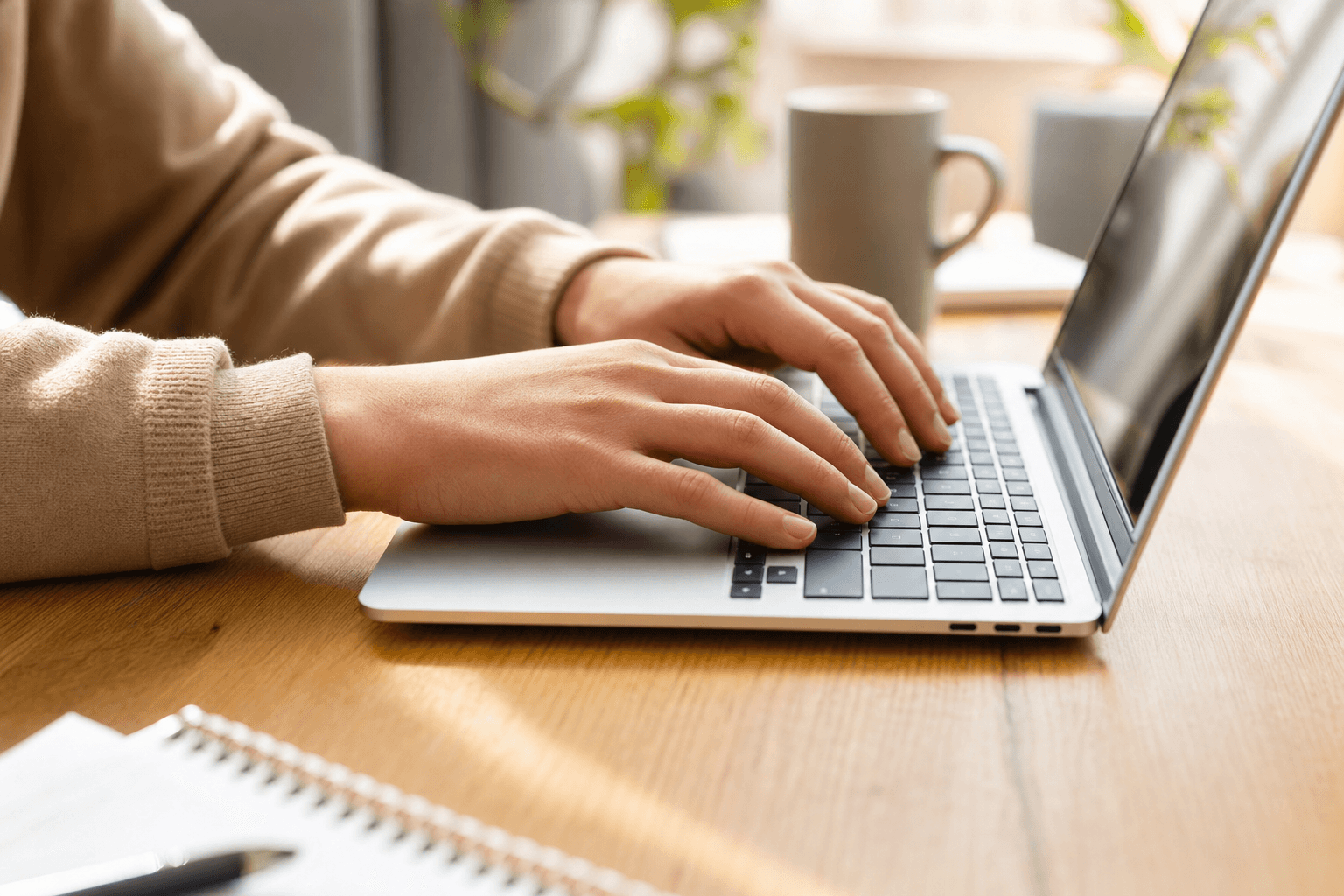 Close-up of a person's hands typing on a laptop keyboard on a bright wooden desk in warm daylight