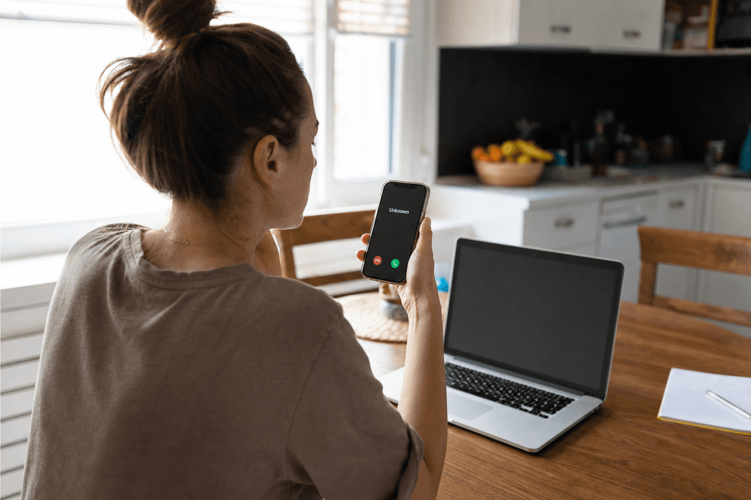 Over-the-shoulder view of a woman at a kitchen table looking at her smartphone screen showing an incoming call from an unknown number, an open laptop in front of her, in daylight
