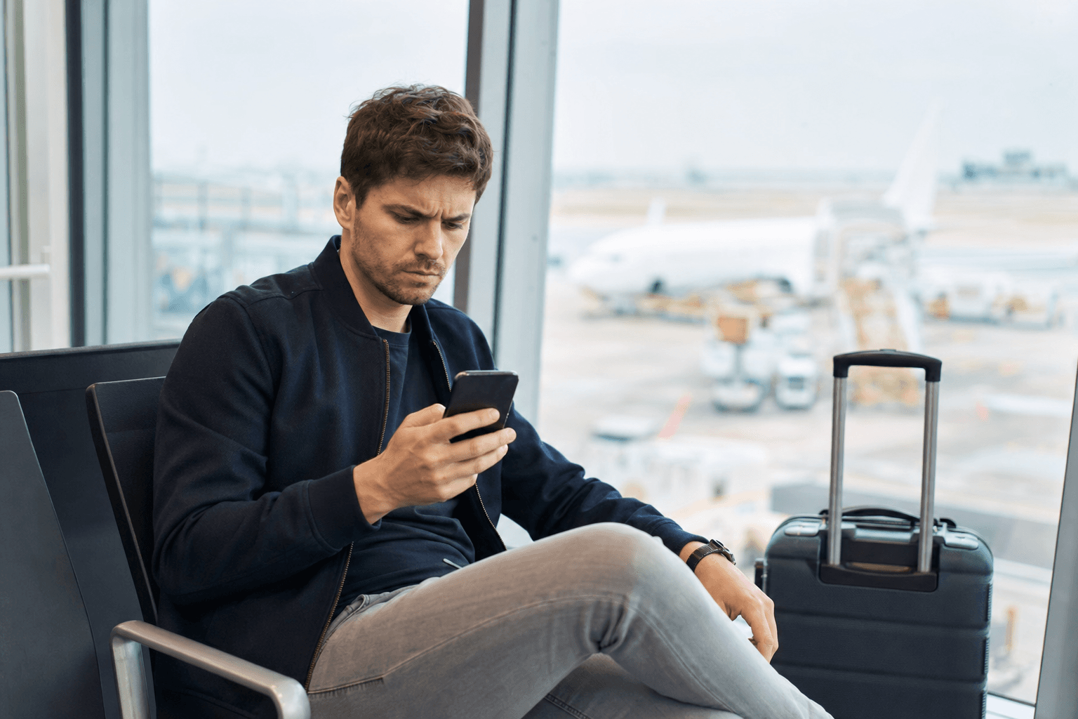 Traveler at an airport gate looking at a phone showing a suspicious flight rebooking text message, suitcase beside them