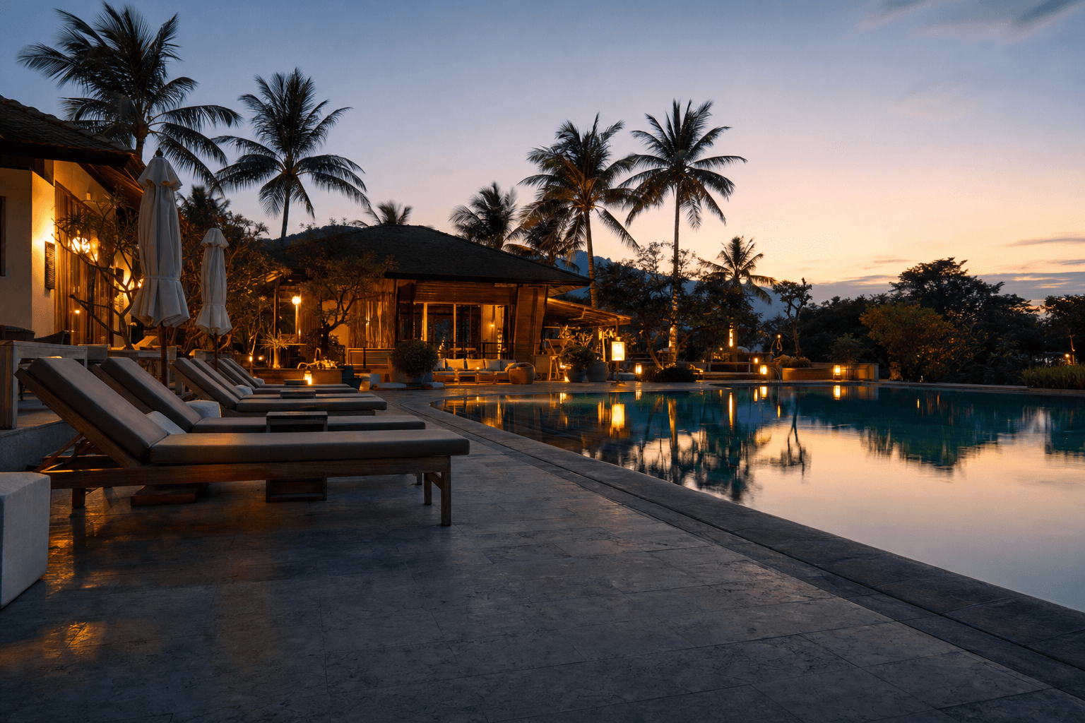 An empty resort swimming pool and lounge chairs at dusk with warm lights coming on